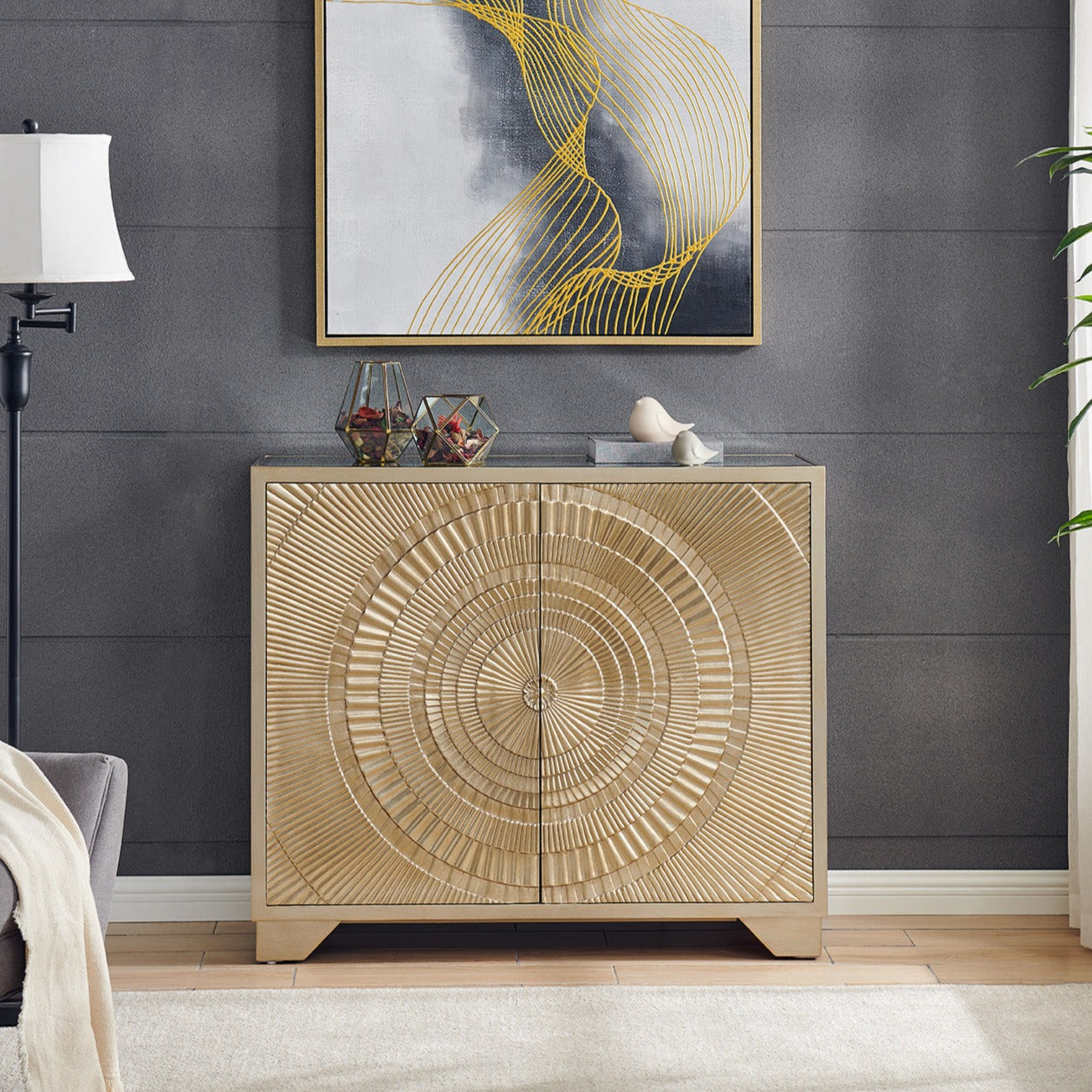 A gold geometric chest with two doors, placed against a wall with a framed abstract painting above it, next to a floor lamp and a plant.