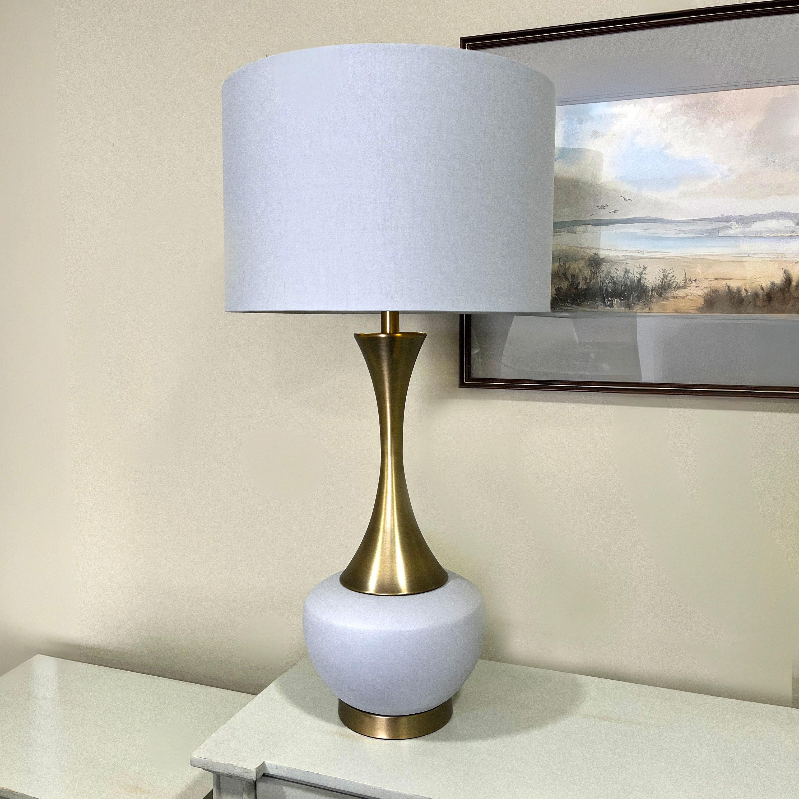 Table lamp with white shade and gold base on a table next to a painting.