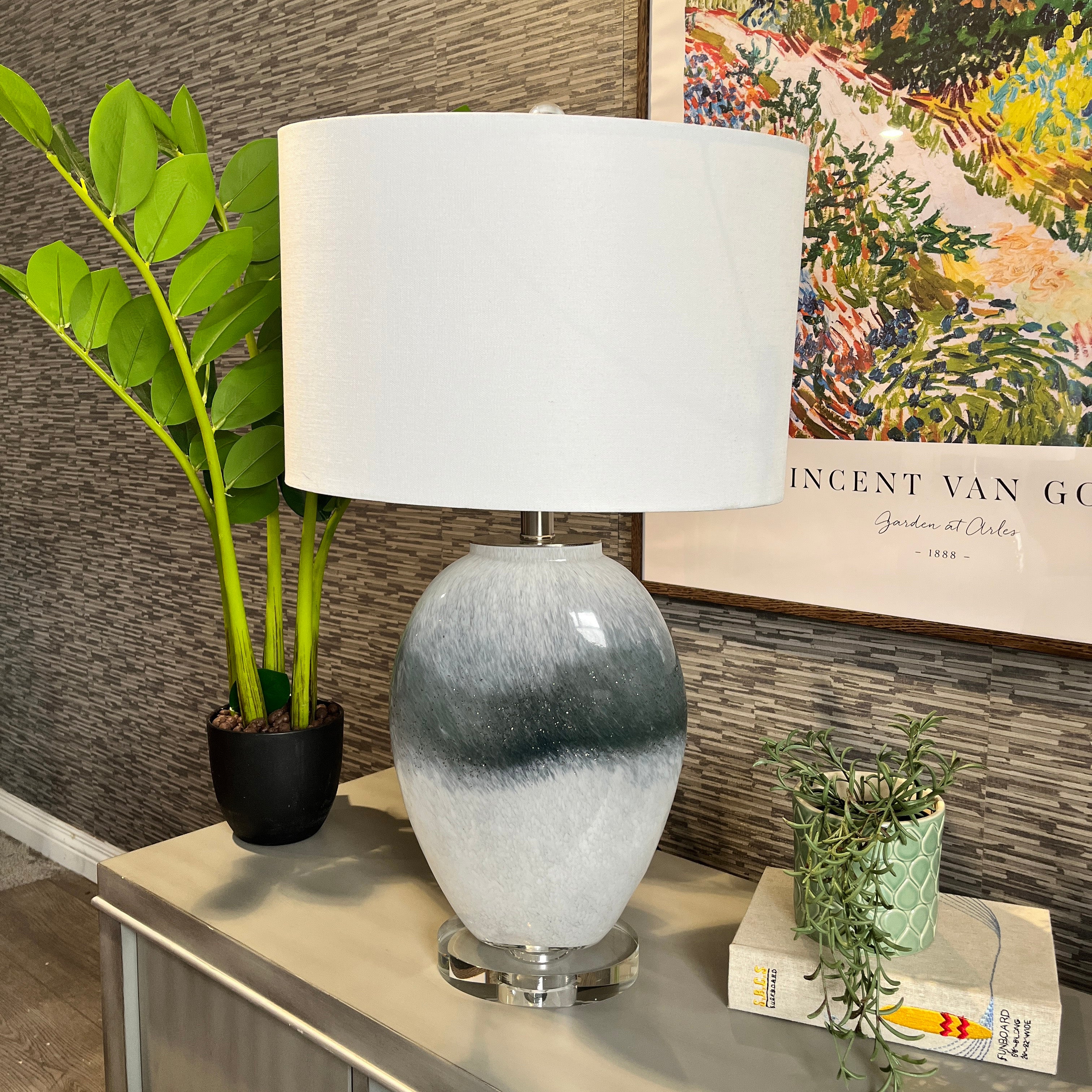 Table lamp with a white shade on a surface next to a plant and books, with a painting in the background.