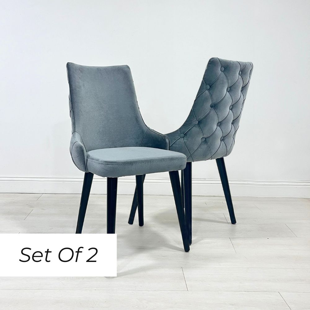 A set of two grey velvet dining chairs with button detailing and a scooped back design, on dark legs, against a white background.