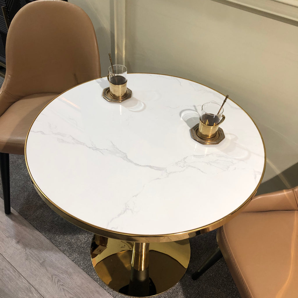 A round dining table with a marble effect top in white, paired with a gold and black metal base, set in an interior with two chairs and some decorative items on the table.