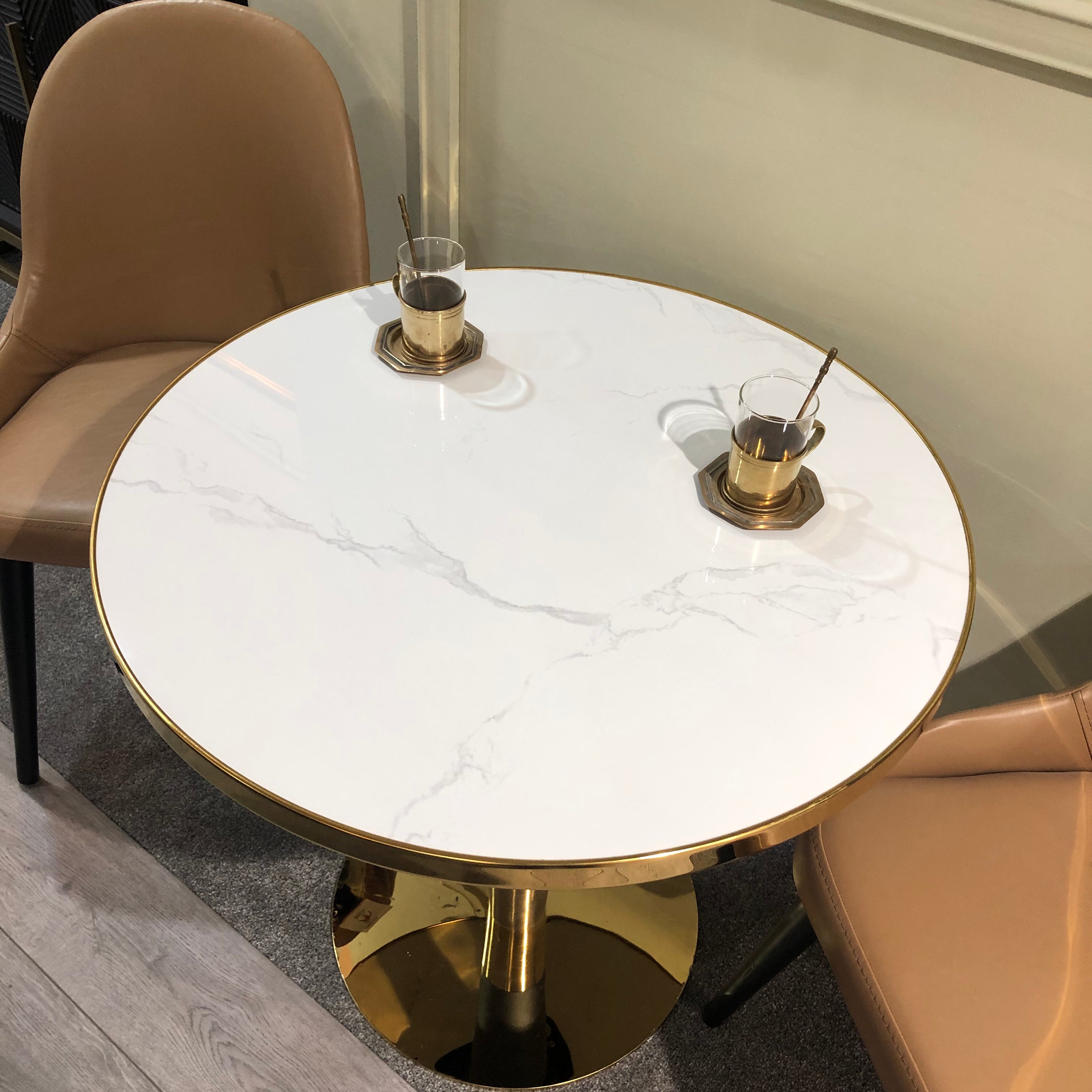 A round dining table with a marble effect top in white, paired with a gold and black metal base, set in an interior with two chairs and some decorative items on the table.