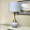Table lamp with white shade and gold base on a table next to a painting.