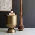 An antique brass storage table with a round shape and a lift-off tabletop, placed beside a wooden column on a grey background.